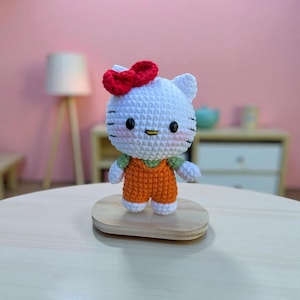 May include: A handmade crochet doll resembling a popular cartoon character. The doll has a white head with black eyes, whiskers, and a red bow. It wears an orange jumpsuit with a green shirt underneath. The doll is displayed on a small wooden stand.