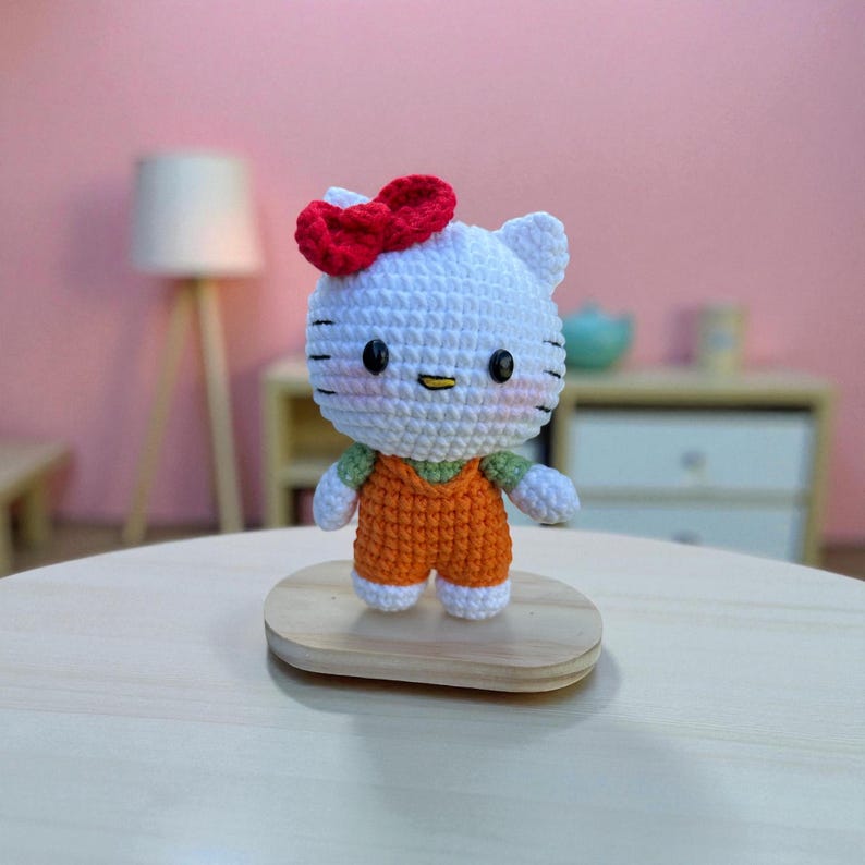 May include: A handmade crochet doll resembling a popular cartoon character. The doll has a white head with black eyes, whiskers, and a red bow. It wears an orange jumpsuit with a green shirt underneath. The doll is displayed on a small wooden stand.