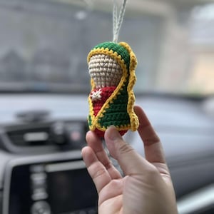 May include: A crocheted figurine of the Virgin Mary, wearing a green and yellow robe, hanging from a rearview mirror in a car.