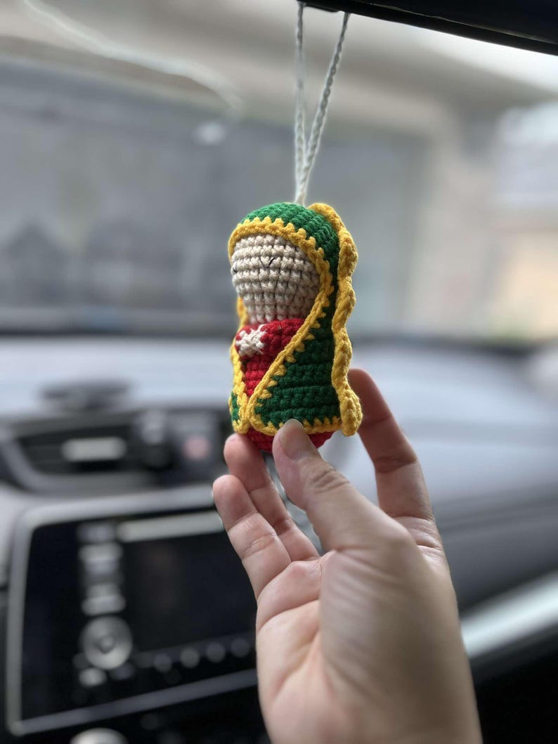 May include: A crocheted figurine of the Virgin Mary, wearing a green and yellow robe, hanging from a rearview mirror in a car.