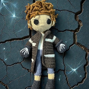 May include: A handmade crochet doll with a brown coat, blue pants, and black button eyes. The doll has brown hair and is wearing a white shirt. The doll's hands have a white arrow design. The doll is a unique, handcrafted item.
