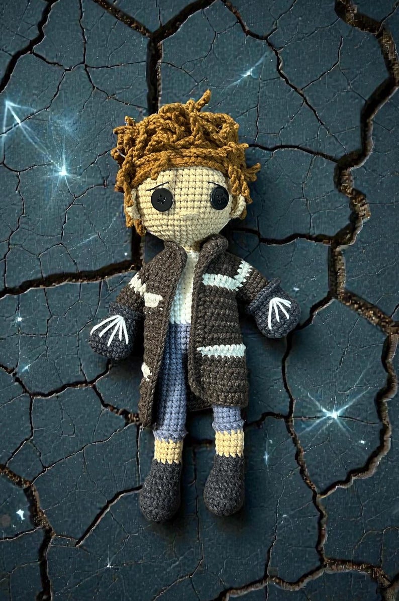 May include: A handmade crochet doll with a brown coat, blue pants, and black button eyes. The doll has brown hair and is wearing a white shirt. The doll's hands have a white arrow design. The doll is a unique, handcrafted item.