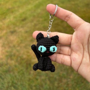 May include: A black crochet cat keychain with bright blue eyes and a silver keyring. The handmade cat has its paw raised and is sitting upright. The cat's ears are perked up, and the keychain is held against a blurred green background.