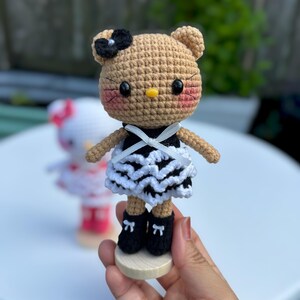 May include: A crocheted doll with a tan head, black bow, and pink cheeks. It wears a black and white dress and black shoes with bows. The doll is on a wooden base. Another doll is visible in the background.