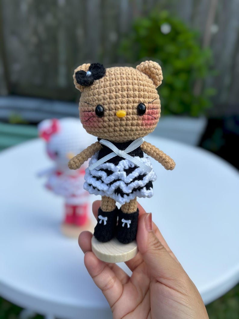 May include: A crocheted doll with a tan head, black bow, and pink cheeks. It wears a black and white dress and black shoes with bows. The doll is on a wooden base. Another doll is visible in the background.