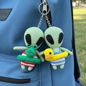 May include: Two crocheted alien keychains hanging from a silver chain. One alien wears a red and white striped floatie and the other wears a yellow and blue floatie. The aliens are green with black eyes.