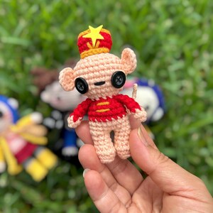 May include: A handmade crochet mouse figurine with a red and yellow crown and a red jacket. The mouse has button eyes and is held against a background of green grass. The figurine is approximately 3 inches tall.