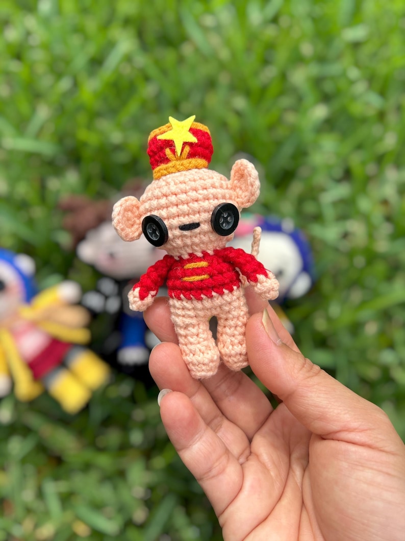 May include: A handmade crochet mouse figurine with a red and yellow crown and a red jacket. The mouse has button eyes and is held against a background of green grass. The figurine is approximately 3 inches tall.