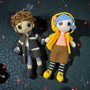May include: Two handmade crochet dolls. One doll has brown hair, a brown jacket, and blue pants. The other doll has blue hair, a yellow jacket, and a brown skirt. Both dolls have black button eyes and are holding hands.