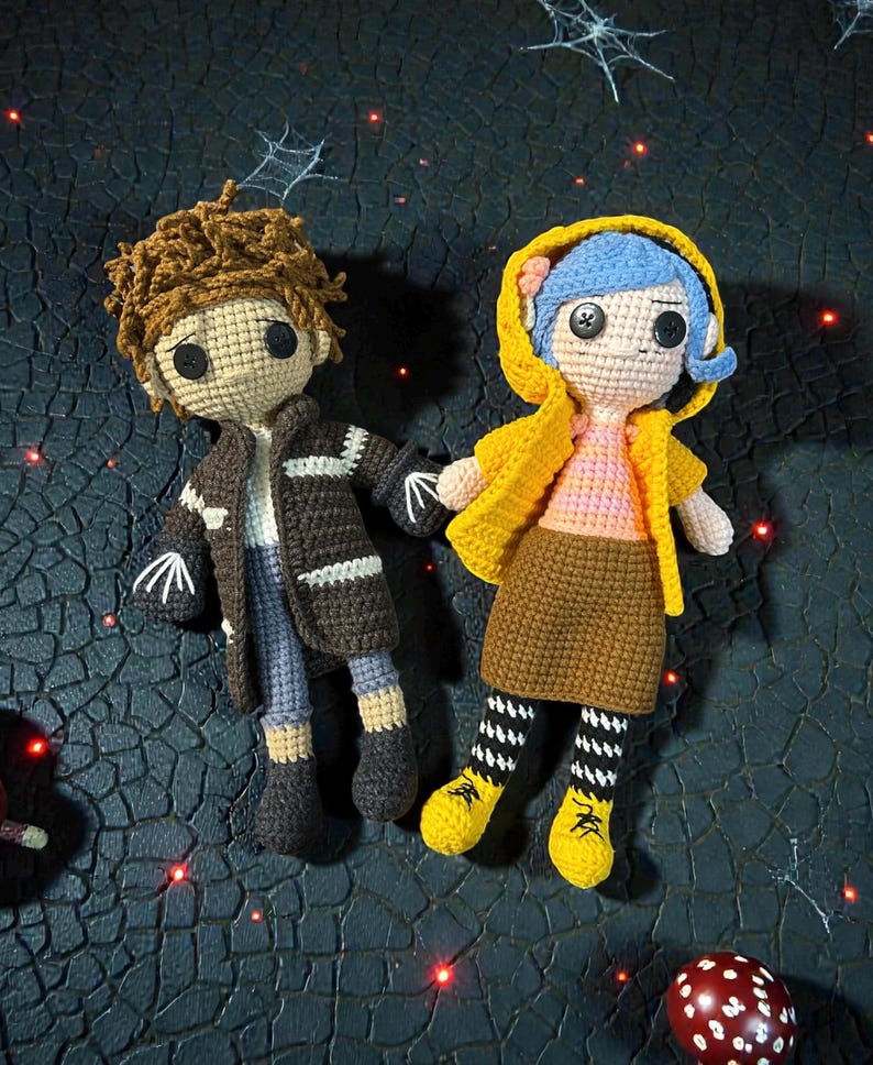 May include: Two handmade crochet dolls. One doll has brown hair, a brown jacket, and blue pants. The other doll has blue hair, a yellow jacket, and a brown skirt. Both dolls have black button eyes and are holding hands.