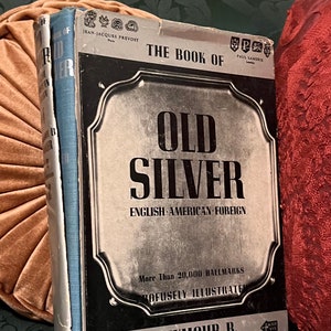 May include: A book titled "The Book of Old Silver: English, American, Foreign" with a silver-colored cover and black text. The book features more than 20,000 hallmarks and is profusely illustrated. The author is Seymour B. Wyler.
