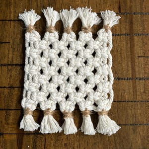 May include: Cream-colored macrame coaster with a square shape. The coaster features a woven pattern with diamond-shaped openings. Each end has a series of white tassels, secured with twine. The coaster is designed to protect surfaces from drink condensation.