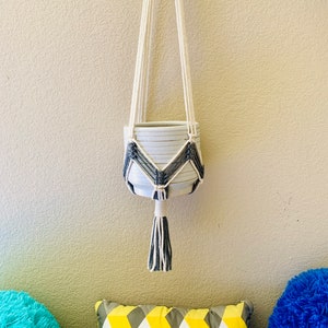 May include: White ceramic planter with a gray geometric pattern hanging from a white macrame plant hanger with a gray tassel.