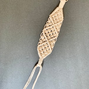 May include: A white macrame keychain with a silver ring at the top. The keychain is made of a thick, knotted cord and has a long, dangling loop at the bottom.