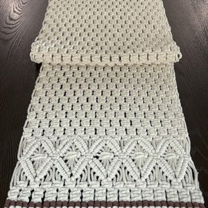 May include: A white crocheted table runner with a brown stripe and fringe. The runner has a geometric pattern.