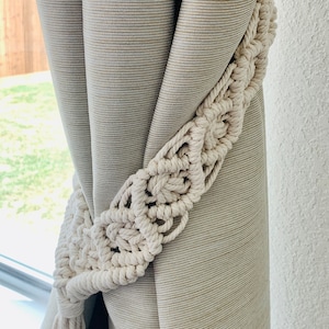 May include: A close-up of a beige curtain with a white macrame curtain tieback. The tieback is a long, flowing piece of macrame with a tassel at the end.