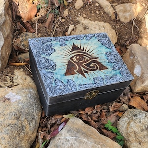 Puede incluir: Una caja de madera negra con una tapa decorativa que presenta un símbolo de ojo dentro de un triángulo, rodeado de líneas radiantes. La tapa tiene un fondo azul claro y un borde texturizado. Un cierre de latón asegura la caja.
