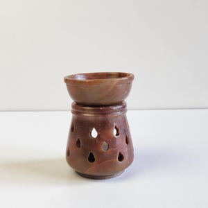 Puede incluir: Un difusor de aceite de piedra tallada marrón. Tiene un cuenco pequeño en la parte superior y una base con cortes en forma de lágrima. El difusor está hecho de piedra marrón rojiza y tiene un acabado liso y pulido. El diseño es simple y elegante.