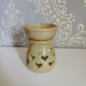 May include: A beige stone oil burner featuring a bowl-shaped top and a cylindrical base with multiple heart-shaped cutouts. The stone has a smooth, mottled texture with subtle variations in color, ranging from light cream to soft tan throughout.