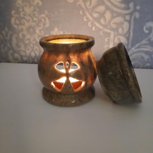 May include: A round, brown, mottled stone oil burner with a cut-out butterfly design on the front. The burner is illuminated from within by a small candle, casting a warm glow through the butterfly shape. A matching stone lid sits beside it.