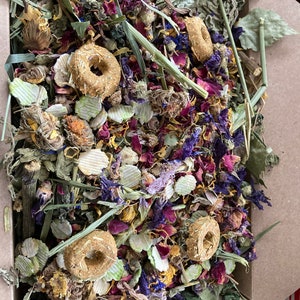 May include: A close-up of a brown cardboard box filled with a mixture of dried herbs, flowers, and small, round, golden-brown treats. The mixture includes dried rose petals, lavender, and other herbs.