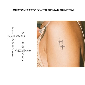 Op de afbeelding: Een close-up van de bovenarm van een persoon met een kleine tatoeage van een kruis gemaakt van Romeinse cijfers. De tatoeage zit op de bovenarm, net boven de elleboog. De tatoeage is zwart-wit en is erg klein.