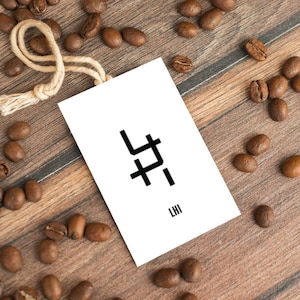 Peut inclure: Une étiquette blanche avec du texte noir et un design graphique simple. L'étiquette est attachée avec une ficelle en ficelle brune. L'étiquette est entourée de grains de café sur une surface en bois. Le texte sur l'étiquette indique "LHI" et le design graphique est un symbole stylisé.
