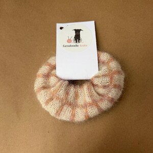 May include: A fuzzy, cream-colored scrunchie featuring a soft peach and pink grid pattern. The hair accessory is attached to a white rectangular display card with a circular logo showing a black dog and the text farndoodle knits.