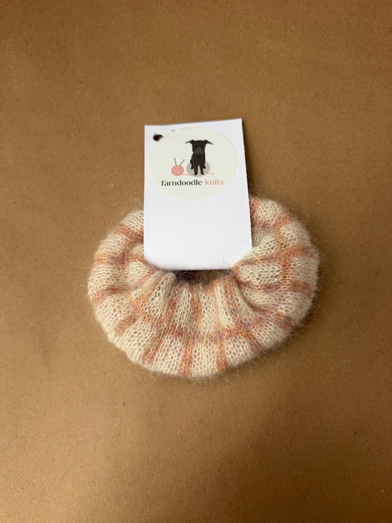 May include: A fuzzy, cream-colored scrunchie featuring a soft peach and pink grid pattern. The hair accessory is attached to a white rectangular display card with a circular logo showing a black dog and the text farndoodle knits.