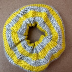 May include: A circular hair scrunchie made of knitted fabric featuring alternating horizontal stripes in bright yellow and light gray. The accessory has a gathered, elasticized construction with a soft, textured yarn surface throughout.