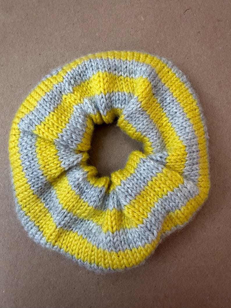 May include: A circular hair scrunchie made of knitted fabric featuring alternating horizontal stripes in bright yellow and light gray. The accessory has a gathered, elasticized construction with a soft, textured yarn surface throughout.