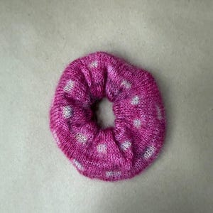 May include: A circular hair scrunchie made of a knitted fabric in a vibrant magenta pink color. The accessory features a repeating pattern of small, light gray or white dots distributed evenly across the textured, soft-looking surface.