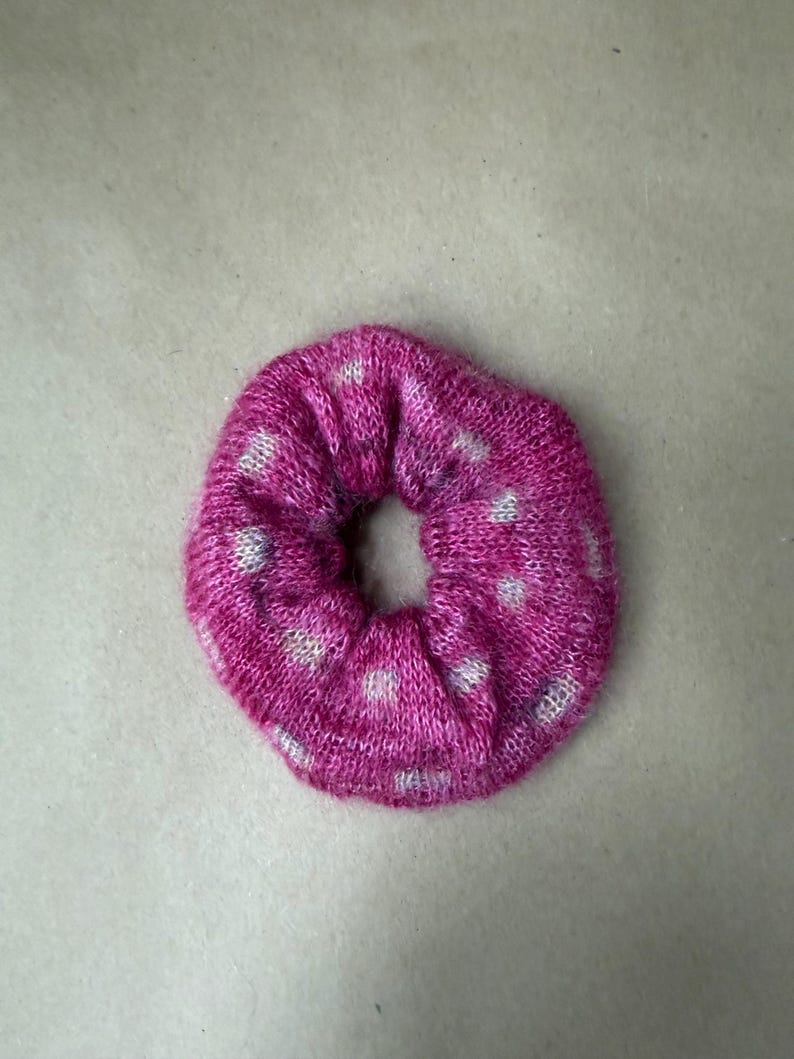 May include: A circular hair scrunchie made of a knitted fabric in a vibrant magenta pink color. The accessory features a repeating pattern of small, light gray or white dots distributed evenly across the textured, soft-looking surface.