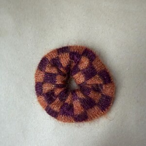 May include: A circular hair scrunchie featuring a fuzzy, knitted texture with a repeating checkered pattern in alternating shades of deep purple and burnt orange. The accessory has a soft, fibrous appearance with a central opening for hair.