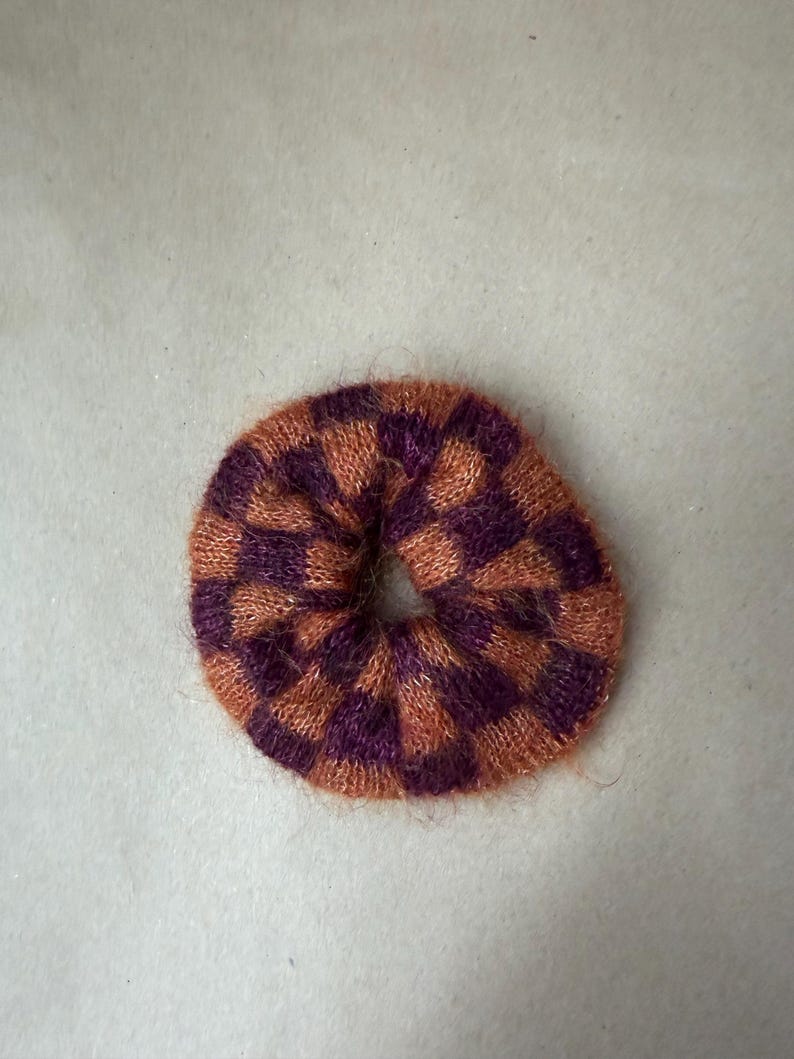 May include: A circular hair scrunchie featuring a fuzzy, knitted texture with a repeating checkered pattern in alternating shades of deep purple and burnt orange. The accessory has a soft, fibrous appearance with a central opening for hair.