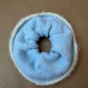 May include: A circular hair scrunchie featuring a light blue knitted center with a gathered texture and a fuzzy, cream-colored knitted border. The accessory has a soft, flexible construction with a central opening, showcasing a two-tone color scheme.