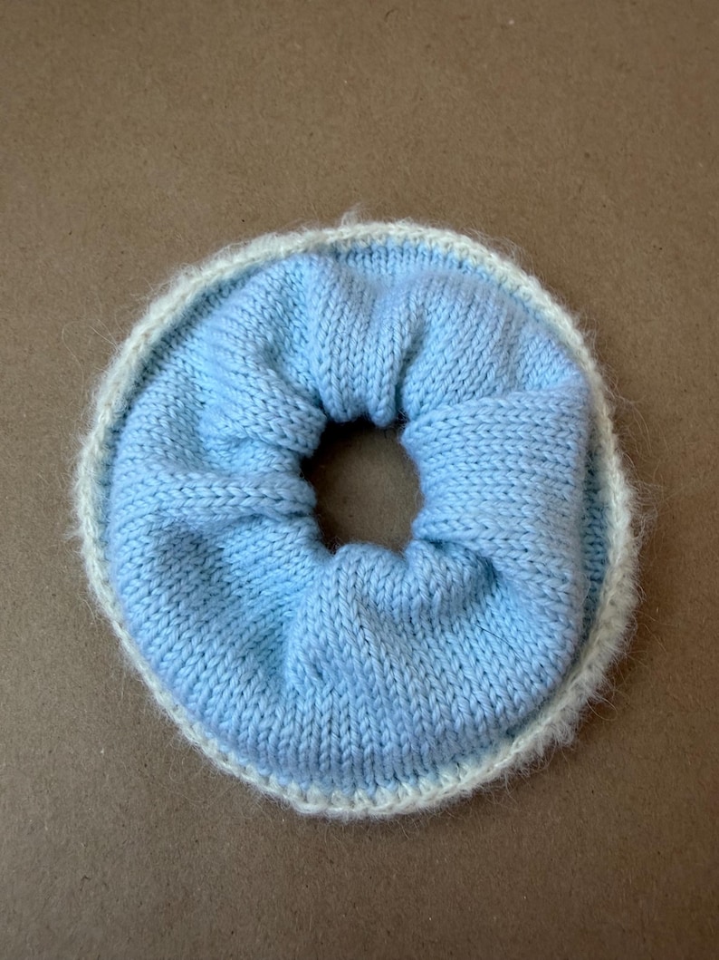 May include: A circular hair scrunchie featuring a light blue knitted center with a gathered texture and a fuzzy, cream-colored knitted border. The accessory has a soft, flexible construction with a central opening, showcasing a two-tone color scheme.