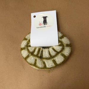May include: A circular, fuzzy hair scrunchie featuring a cream and olive green grid pattern. The scrunchie is attached to a white rectangular display card with a circular logo showing a black dog, a pink ball of yarn, and the text farndoodle knits.