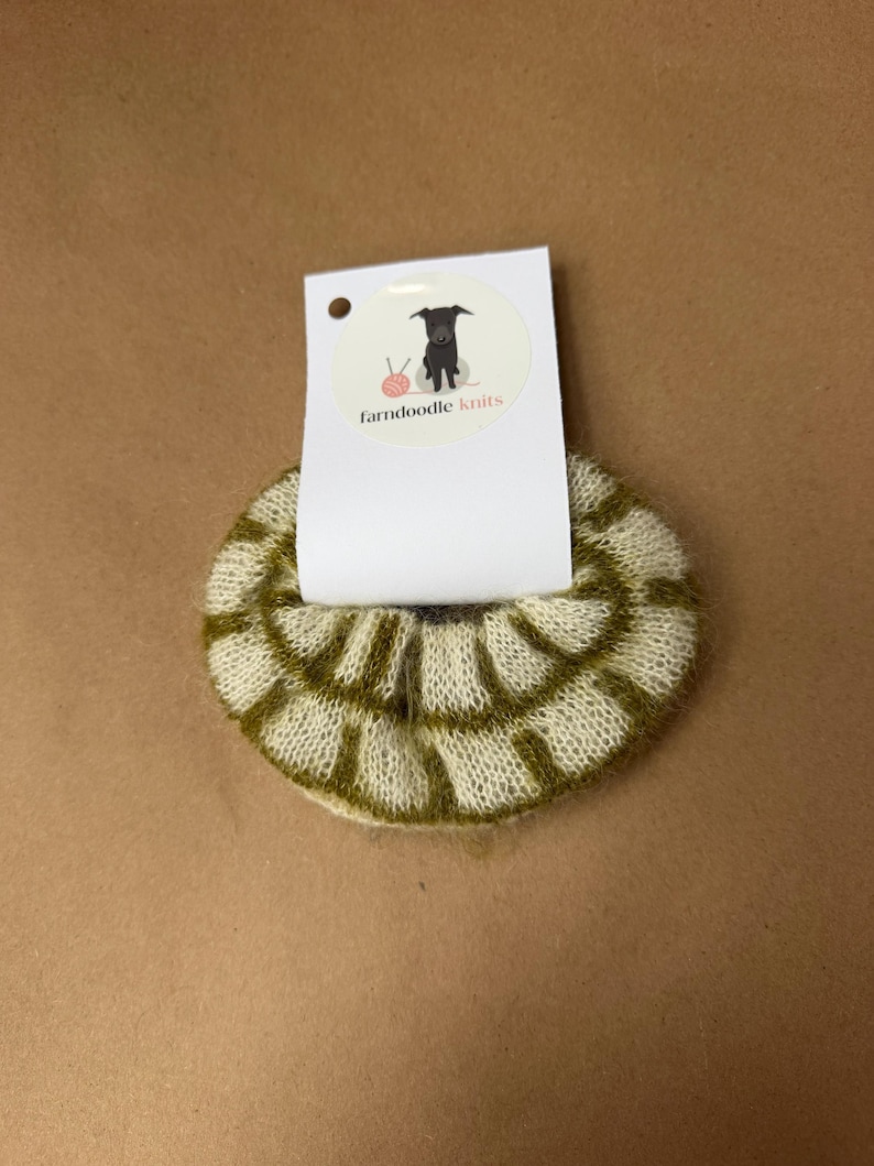 May include: A circular, fuzzy hair scrunchie featuring a cream and olive green grid pattern. The scrunchie is attached to a white rectangular display card with a circular logo showing a black dog, a pink ball of yarn, and the text farndoodle knits.