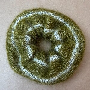 May include: A circular hair scrunchie made of fuzzy, olive green yarn with two concentric white stripes. The accessory features a soft, brushed texture and a gathered, elasticized construction that creates a ruffled, voluminous appearance.