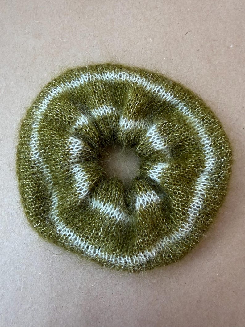 May include: A circular hair scrunchie made of fuzzy, olive green yarn with two concentric white stripes. The accessory features a soft, brushed texture and a gathered, elasticized construction that creates a ruffled, voluminous appearance.