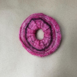 May include: A circular hair scrunchie made of soft, knitted yarn in shades of vibrant magenta, bright pink, and deep purple. The fabric is gathered into a ruffled, elasticized ring shape with a visible, textured knit pattern throughout the accessory.
