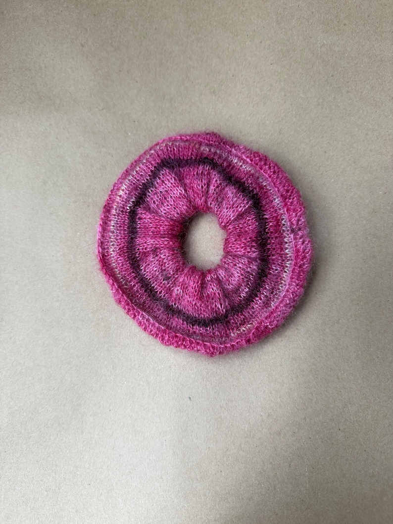 May include: A circular hair scrunchie made of soft, knitted yarn in shades of vibrant magenta, bright pink, and deep purple. The fabric is gathered into a ruffled, elasticized ring shape with a visible, textured knit pattern throughout the accessory.