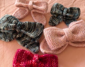 May include: A collection of hand-knitted bow hair accessories in various colors. The bows are in shades of pink, blue, and red. The knitted texture is visible, and the bows are arranged on a pink surface. These bows are perfect for adding a touch of handmade charm to any hairstyle.