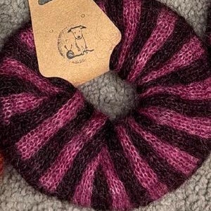 May include: A round, knitted pumpkin-shaped object featuring alternating dark purple and magenta stripes with a grey center stem. A tan cardboard tag is attached to the top, displaying a line-art illustration of a dog sitting next to a ball of yarn.