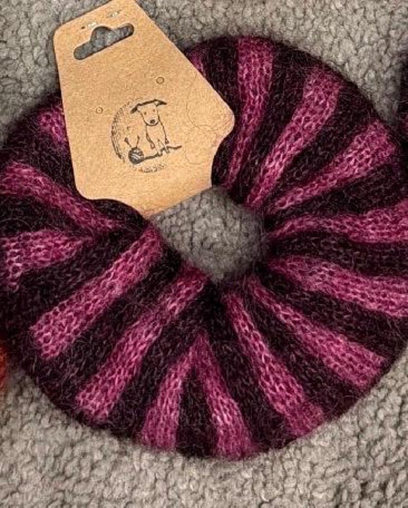 May include: A round, knitted pumpkin-shaped object featuring alternating dark purple and magenta stripes with a grey center stem. A tan cardboard tag is attached to the top, displaying a line-art illustration of a dog sitting next to a ball of yarn.