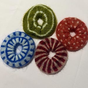 May include: Four fuzzy circular hair scrunchies with distinct patterns. One is olive green with white rings, one is bright blue with white radial stripes, one is dark red with white polka dots, and one is dark red with light pink radial stripes.