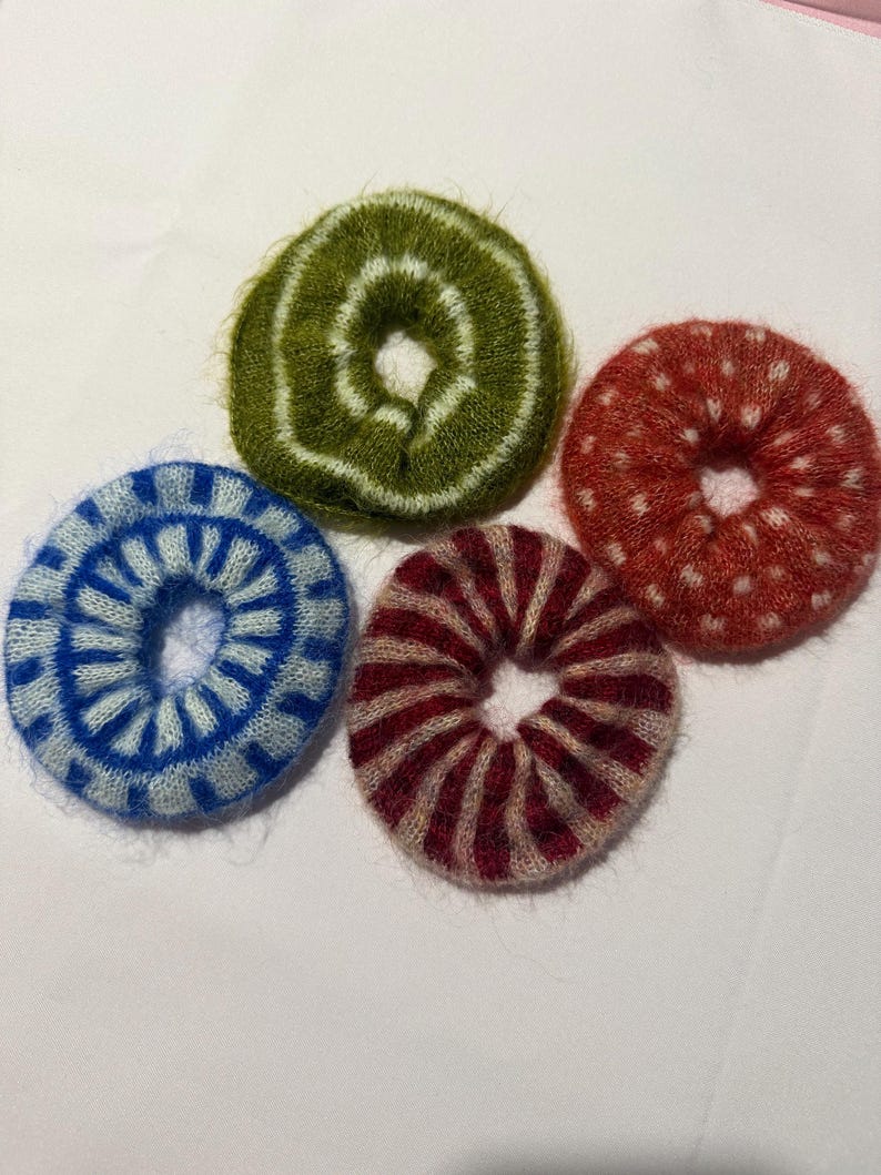 May include: Four fuzzy circular hair scrunchies with distinct patterns. One is olive green with white rings, one is bright blue with white radial stripes, one is dark red with white polka dots, and one is dark red with light pink radial stripes.