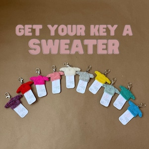 Peut inclure: Une collection de porte-clés en forme de mini-pulls de différentes couleurs, dont le rouge, le rose et le bleu. Chaque porte-clés est doté d'un fermoir en métal et d'une petite étiquette. Le texte "GET YOUR KEY A SWEATER" est affiché au-dessus.