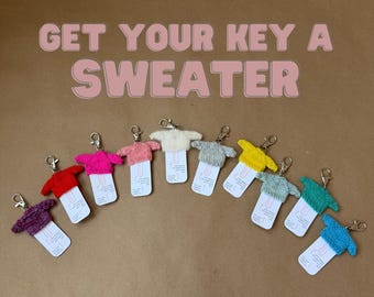 May include: A collection of keychains shaped like miniature sweaters in various colors, including red, pink, and blue. Each keychain has a metal clasp and a small tag. The text "GET YOUR KEY A SWEATER" is displayed above the keychains.